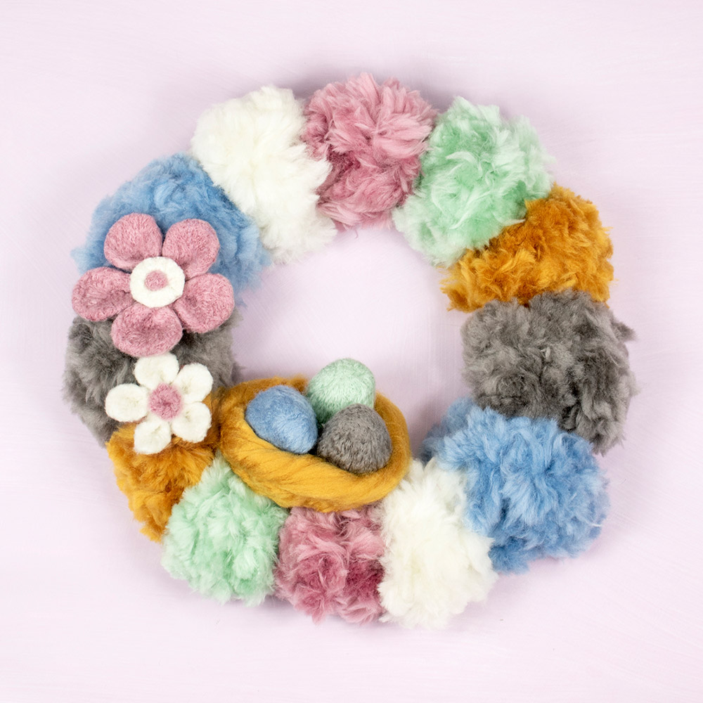 pastel coloured yarns decorate a circle spring pom pom wreath with easter eggs in basket needle felted decoration 